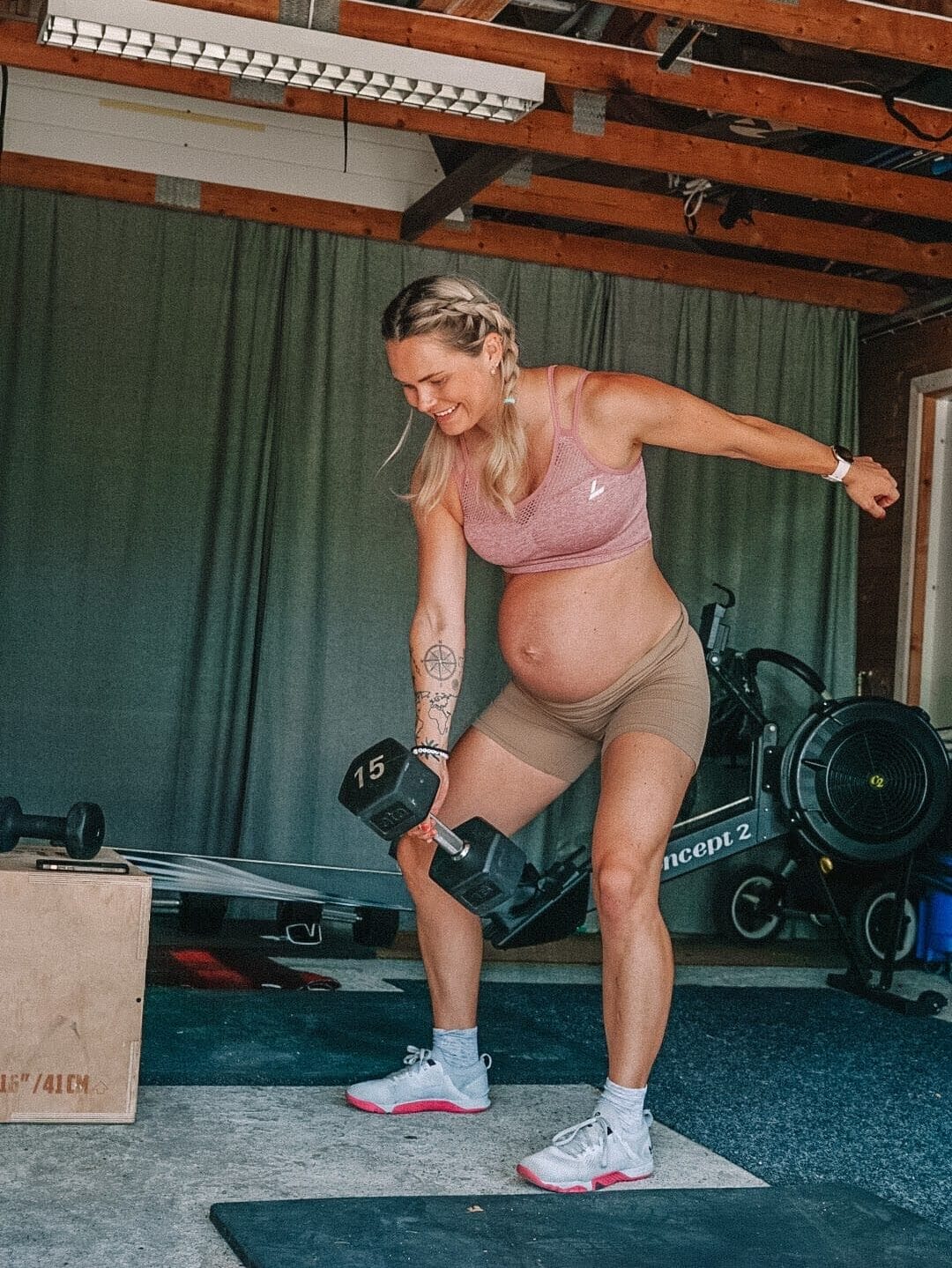 Gravid kvinne som står med bøyde knær og løfter en treningsmanual på 15 kg med høyre hånd.